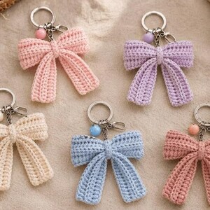 Può includere: Cinque portachiavi a forma di fiocco all'uncinetto in colori pastello, tra cui rosa chiaro, lavanda, crema, azzurro e rosa antico. Ogni fiocco è fissato a un anello in metallo argentato con una piccola perlina abbinata e un moschettone.