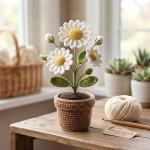 Puede incluir: Una planta de margaritas tejida a ganchillo en una maceta marrón con pétalos blancos y centros amarillos, con tallos y hojas verdes. Una pequeña etiqueta de papel sobre la mesa dice "CROCHET PATTERN - DAISY POT". La planta está hecha de hilo con puntos visibles.