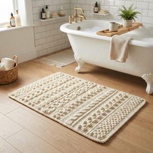 May include: A rectangular cream-colored bath mat featuring a textured knit pattern with alternating rows of raised bobbles and braided cable stitches. The mat is crafted from thick yarn and lies flat on a light wood floor in front of a white bathtub.