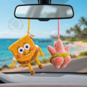 Op de afbeelding: Twee gehaakte hangende ornamenten aan de achteruitkijkspiegel van een auto. De ene is een geel vierkant figuur met blauwe ogen en een bruine onderkant, de andere is een roze stervormig figuur met een groene broek. Beide zijn gemaakt van garen.