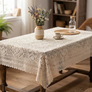 May include: A rectangular beige crochet tablecloth featuring an intricate openwork pattern with geometric and floral motifs. The fabric drapes over the edges of a wooden table, showcasing a scalloped border design and a detailed lace-like texture.
