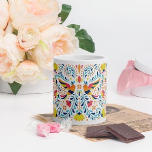 May include: White ceramic mug featuring a colourful floral and bird design. The design incorporates blue, yellow, orange, red, and green hues. The mug is positioned on a table with a newspaper and sweets.