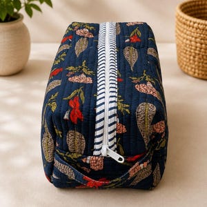 May include: A rectangular quilted toiletry bag in dark blue fabric featuring a repeating pattern of leaves and flowers in shades of red, pink, and beige. The bag includes a white zipper with a blue and white striped trim and a handle.