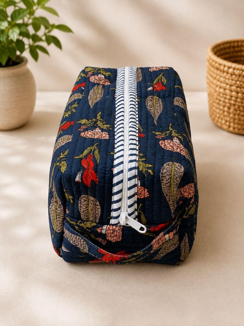 May include: A rectangular quilted toiletry bag in dark blue fabric featuring a repeating pattern of leaves and flowers in shades of red, pink, and beige. The bag includes a white zipper with a blue and white striped trim and a handle.
