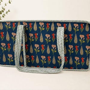 May include: A rectangular quilted tote bag in deep blue featuring a repeating pattern of red flowers and beige teardrop shapes. The bag has two striped handles and matching striped piping along the edges, providing a structured appearance.