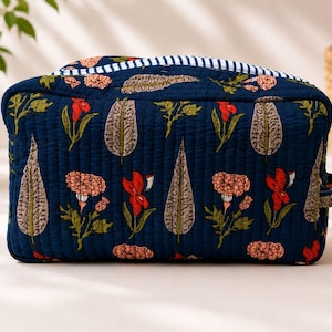 May include: A rectangular quilted toiletry bag in dark blue fabric featuring a repeating pattern of orange flowers and grey leaves. The top edge is accented with a white and blue striped trim, and a small fabric loop is attached to the side.