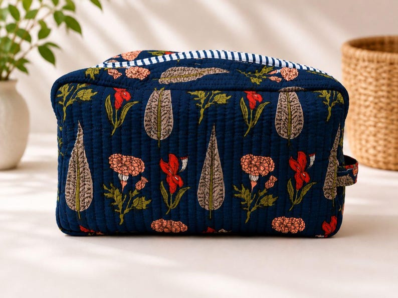 May include: A rectangular quilted toiletry bag in dark blue fabric featuring a repeating pattern of orange flowers and grey leaves. The top edge is accented with a white and blue striped trim, and a small fabric loop is attached to the side.