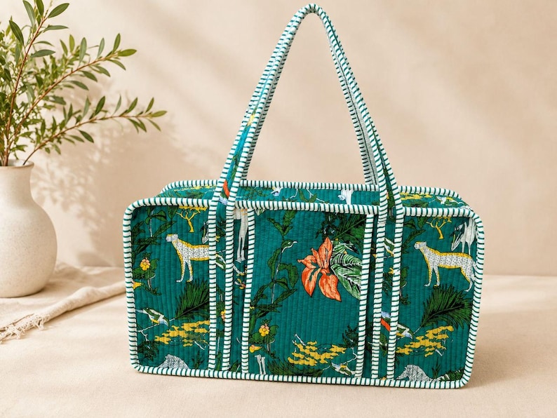 May include: A rectangular quilted tote bag in deep teal featuring a repeating pattern of cheetahs, tropical foliage, and orange flowers. The bag has structured sides and handles with a white and teal striped trim along all edges.