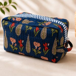 May include: A rectangular quilted toiletry bag in deep navy blue fabric featuring a repeating floral and leaf pattern in shades of red, pink, and beige. The bag includes a white and navy blue striped zipper closure and a small side handle.