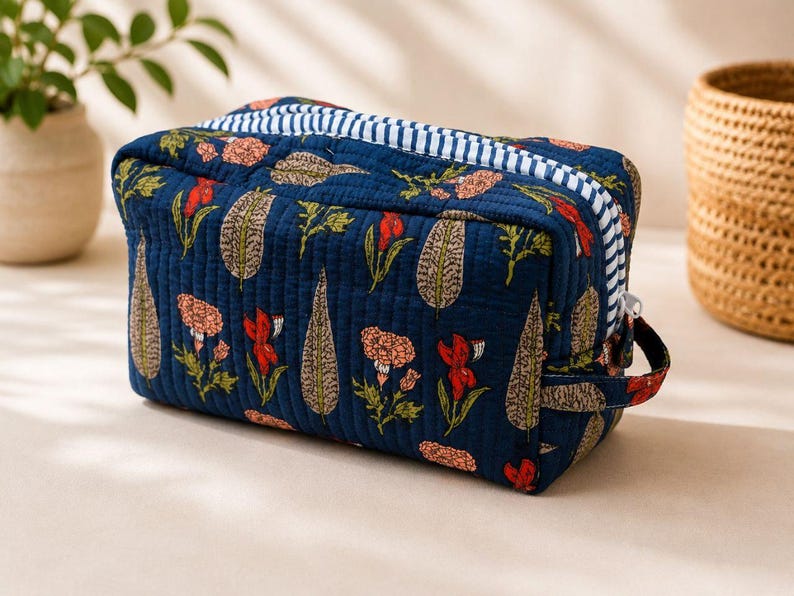 May include: A rectangular quilted toiletry bag in deep navy blue fabric featuring a repeating floral and leaf pattern in shades of red, pink, and beige. The bag includes a white and navy blue striped zipper closure and a small side handle.