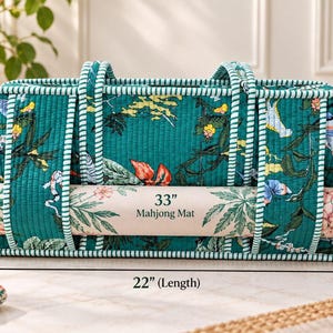 May include: A teal quilted fabric bag with a floral and bird pattern, featuring striped handles and trim. The bag holds a rolled 33-inch Mahjong mat. Dimensions are 22 inches in length, 11 inches in height, and 6 inches in width.