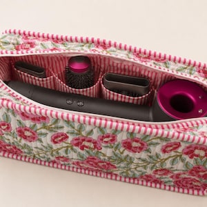 May include: A rectangular quilted storage case featuring a pink and green floral pattern on a white background with pink and white striped trim. The interior contains a dark gray hair dryer with a magenta circular head and three hair styling attachments.