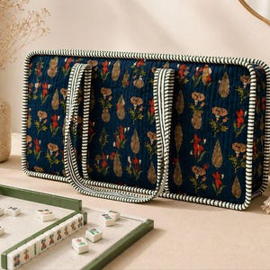 May include: A rectangular quilted tote bag in dark blue fabric featuring a repeating floral and paisley print in red, orange, and green. The bag has two handles and edges trimmed with a white and dark green striped fabric piping.