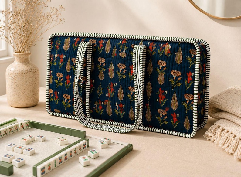 May include: A rectangular quilted tote bag in dark blue fabric featuring a repeating floral and paisley print in red, orange, and green. The bag has two handles and edges trimmed with a white and dark green striped fabric piping.