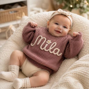 Könnte beinhalten: Ein altrosa langärmeliger Strickpullover mit dem in weißem Garn aufgestickten Namen Mila auf der Brust und einem kleinen Gänseblümchen-Akzent. Dazu gehören cremefarbene Strickstrümpfe und ein passendes cremefarbenes Stirnband mit einer Schleife.