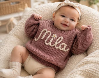 May include: A dusty rose knitted long-sleeve sweater featuring the name Mila embroidered in white yarn across the chest with a small daisy accent. The sweater is paired with cream-colored knit socks and a matching cream-colored headband with a bow.
