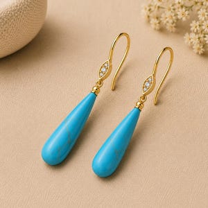 May include: A pair of gold drop earrings featuring smooth, teardrop-shaped turquoise stones. Each earring has a curved gold hook and a small, marquise-shaped accent piece set with three tiny, clear, sparkling stones above the turquoise drop.