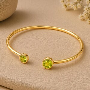 May include: A gold-toned open cuff bracelet featuring two round, faceted lime green stones set in circular bezels at each end of the thin, polished metal band. The design is simple and circular, showcasing the vibrant green gems clearly.