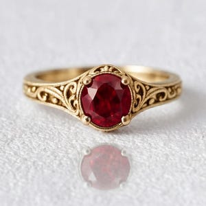 May include: A gold ring featuring a round, deep red gemstone set in the centre. The band is crafted with intricate, swirling metal filigree patterns that extend from the central setting along the sides of the ring, creating a detailed texture.