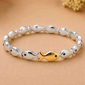 Könnte beinhalten: Ein kreisförmiges Armband aus sich wiederholenden silberfarbenen fischförmigen Gliedern, die jeweils einen kleinen schwarzen Punkt als Auge aufweisen. Ein einzelnes goldfarbenes fischförmiges Glied ist markant zwischen den silbernen Gliedern platziert.