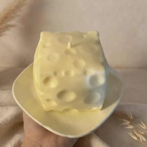 Könnte beinhalten: Eine blassgelbe, würfelförmige Kerze, die einem Stück Schweizer Käse nachempfunden ist und zahlreiche kreisförmige Vertiefungen auf ihrer Oberfläche aufweist. Die Kerze steht auf einer passenden blassgelben, quadratischen Untertasse.