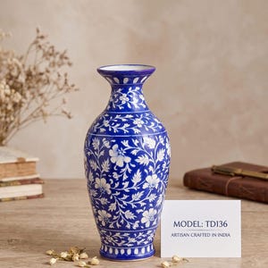 以下が含まれることがあります： 青と白の陶器製の花瓶で、蔓や葉が描かれた詳細な花柄が特徴です。花瓶の前には白い長方形のカードが置かれており、MODEL: TD136 ARTISAN CRAFTED IN INDIA という文字が記されています。花瓶は縁と底が広がった形状をしています。