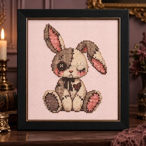 May include: A cross-stitch embroidery of a seated rabbit with a patchwork appearance, featuring one button eye, stitched details, and a small heart on its chest. The rabbit is rendered in shades of brown, cream, and pink, set against a light pink fabric background and enclosed in a black frame with a thin gold inner border.
