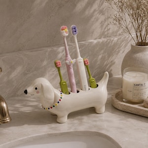 Peut inclure: Un porte-brosse à dents en céramique blanche en forme de teckel avec un collier perlé coloré. Le support présente quatre ouvertures circulaires sur le dos, contenant actuellement deux brosses à dents vertes, une rose et une blanche.