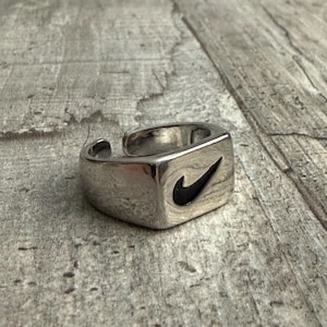 Può includere: Un anello in metallo color argento con una faccia rettangolare caratterizzata da un logo Nike swoosh nero inciso. La fascia è aperta sul retro, consentendo una regolazione della misura, e presenta una finitura lucida su tutta la superficie.
