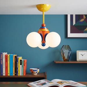 May include: A ceiling light fixture featuring a yellow canopy, a central stem, and a multi-colored base in orange, blue, and red. The fixture includes three spherical white glass globes arranged around the central structure to provide illumination.