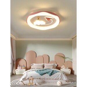 May include: A circular flush mount ceiling light fixture with a pink outer rim and a white illuminated centre. The design features a small gold sphere, a white cloud shape, and a pink paper aeroplane silhouette integrated into the light panel.