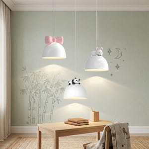 Può includere: Tre lampade a sospensione bianche a forma di cupola che pendono dal soffitto. Una lampada presenta un grande fiocco rosa sulla parte superiore, un'altra ha una piccola figura di panda e la terza è sormontata da un coniglietto bianco.