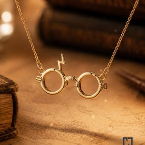 Op de afbeelding: Een goudkleurige ketting met een hanger in de vorm van een ronde bril, voorzien van een bliksemschichtsymbool aan de bovenkant van het linker montuur. De hanger hangt aan een fijne goudkleurige ketting met een gepolijste afwerking.