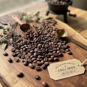 Könnte beinhalten: Ein Haufen dunkel gerösteter Kaffeebohnen auf einem Holzbrett mit einer Kupferschaufel, einem goldenen Löffel und einem beigen Papieranhänger mit dem Text: ARTISAN HAND ROASTED - SINGLE ORIGIN - LIMITED BATCH. Eine kleine schwarze Kaffeemühle ist zu sehen.