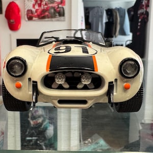 May include: A white scale model sports car featuring a black and orange racing stripe down the center, the number 91 in a black circle on the hood, two circular headlights, two small orange signal lights, and a visible radiator fan assembly.