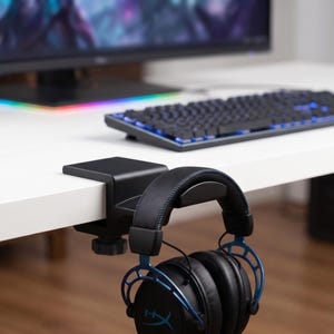 Könnte beinhalten: Ein schwarzer Kopfhörerhalter aus Metall, der an der Kante eines weißen Schreibtisches befestigt ist und einen schwarzen Over-Ear-Kopfhörer mit blauen Akzenten und dem HyperX-Logo auf den Ohrmuscheln trägt, inklusive eines Schraubmechanismus.