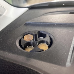 May include: A black, circular, 3D-printed coin organizer insert featuring four distinct compartments arranged around a central post. The organizer is fitted into a circular dashboard recess and contains several metallic euro coins.
