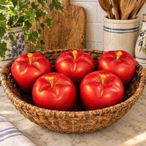 May include: A round, dark brown woven basket containing five smooth, bright red artificial apples. Each apple features a small, light-colored stem at the top. The basket has a textured, rustic appearance and is filled with these round, glossy red fruits.