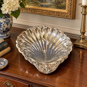 May include: A polished silver serving dish shaped like a large scallop shell. The interior features deep, radiating fluted grooves, while the base is decorated with intricate embossed scrollwork and a small shell motif at the bottom edge.