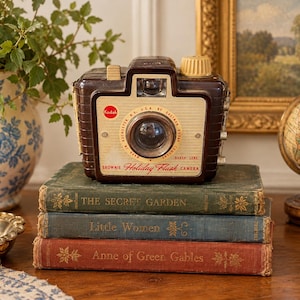 May include: A vintage brown Kodak Brownie Holiday Flash camera with a cream-colored faceplate, featuring the text Kodak, Made in Rochester, N.Y., U.S.A. by Eastman Kodak Company, and Brownie Holiday Flash Camera, resting on a stack of three books.