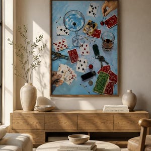Pode incluir: Uma impressão de arte de parede emoldurada apresentando uma superfície azul-clara espalhada com cartas de baralho, uma taça de martini, um copo de uísque, um conjunto de chaves, um isqueiro preto, uma nota de um dólar e duas cerejas seguradas por uma mão.
