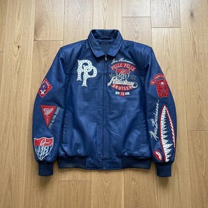 Könnte beinhalten: Eine blaue Lederjacke mit Frontreißverschluss, Kragen und gerippten Bündchen. Die Jacke ist mit verschiedenen gestickten Aufnähern verziert, darunter ein großes weißes PP-Logo, die Schriftzüge Pelle Pelle, Bruiser und Marc Buchanan sowie Haifischzähne.