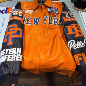Puede incluir: Una chaqueta de cuero naranja con mangas negras que presenta la marca New York Knicks y Pelle Pelle. El frente muestra New York en letras azules, mientras que las mangas incluyen parches con el texto Eastern Conference, Pelle Pelle y NBA.