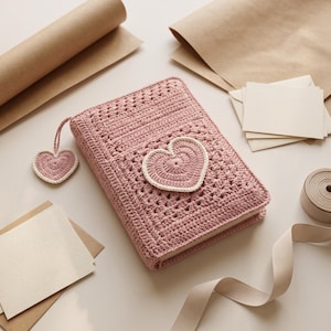 May include: A dusty rose crochet book cover featuring a textured stitch pattern and a prominent heart-shaped applique on the front. A matching heart-shaped crochet charm hangs from a thin cord attached to the spine of the rectangular book cover.