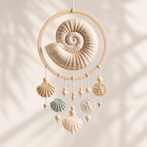 May include: A wall hanging featuring a large, central crocheted nautilus shell inside a circular wooden hoop. Below the hoop, several smaller crocheted shells and sand dollars hang from strings, accented with light wooden and small blue beads.
