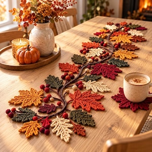 May include: A crochet table runner featuring a vine design with autumn-colored leaves in shades of red, orange, yellow, and green, accented with small crocheted berries. A matching leaf-shaped coaster sits under a ceramic mug of coffee.
