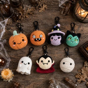 May include: A collection of seven crocheted Halloween-themed keychains featuring a pumpkin, an orange creature, a witch, a green monster, a white ghost, a vampire with a red cape, and a mummy, each attached to a black metal lobster clasp.
