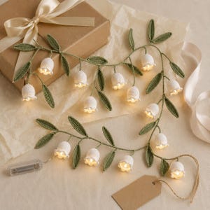May include: A string of crochet lily of the valley flowers featuring white bell-shaped blossoms and green leaves, illuminated by small internal lights. The garland includes a clear plastic battery pack and a brown paper tag attached to the end.