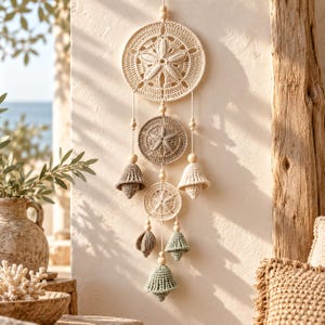 May include: A wall hanging featuring three circular crochet mandalas with sand dollar patterns, accented by wooden beads and five bell-shaped crochet ornaments in shades of cream, beige, and sage green, suspended vertically by thin white cords.