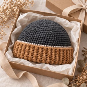 May include: A two-toned crochet beanie featuring a dark charcoal gray crown and a tan ribbed brim. The two sections are separated by a thin, white horizontal stripe. The hat is presented inside a square cardboard gift box lined with white tissue paper.